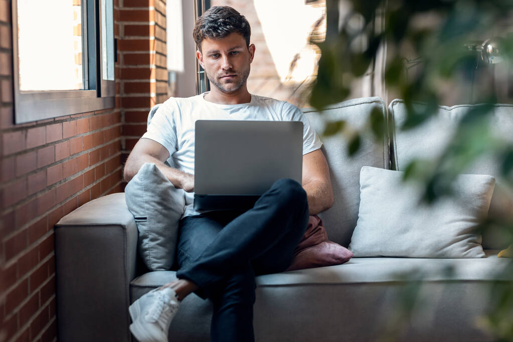 Aufnahme eines intelligenten Mannes, der mit ihrem Laptop arbeitet, während er zu Hause auf einer Couch sitzt - Foto, Bild