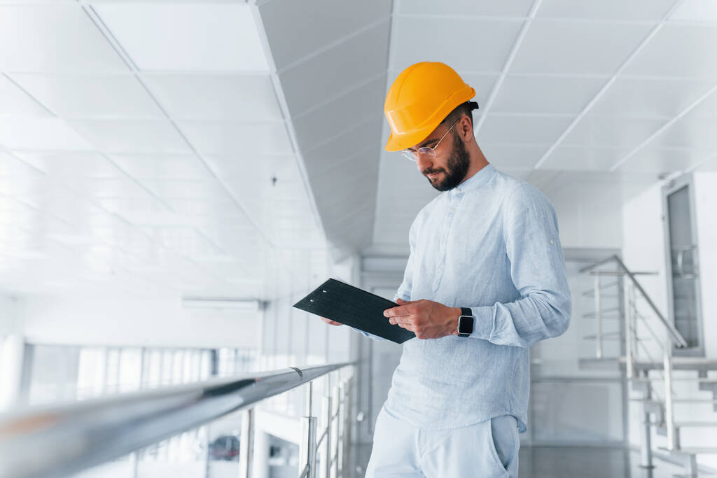 Holding notepad. Engineer in white clothes and orange protective hard hat standing and working indoors. - Foto, afbeelding