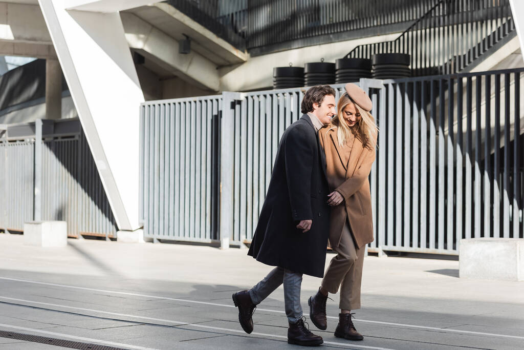 longitud completa de elegante y feliz pareja en abrigos caminando por la calle urbana - Foto, Imagen