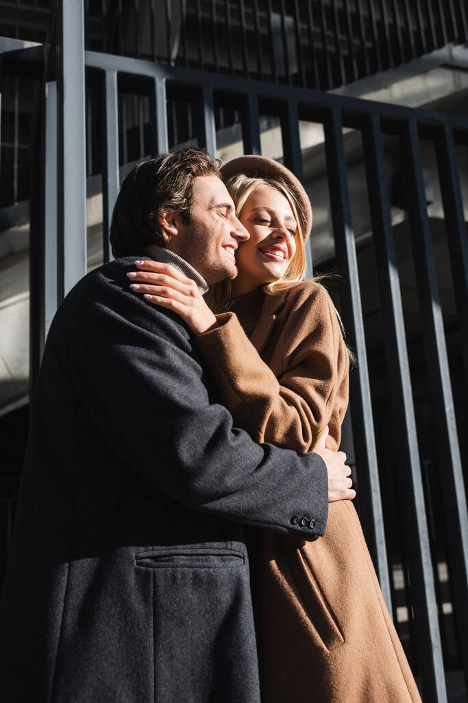 coppia soddisfatta in cappotti abbracciando con gli occhi chiusi vicino alla recinzione - Foto, immagini