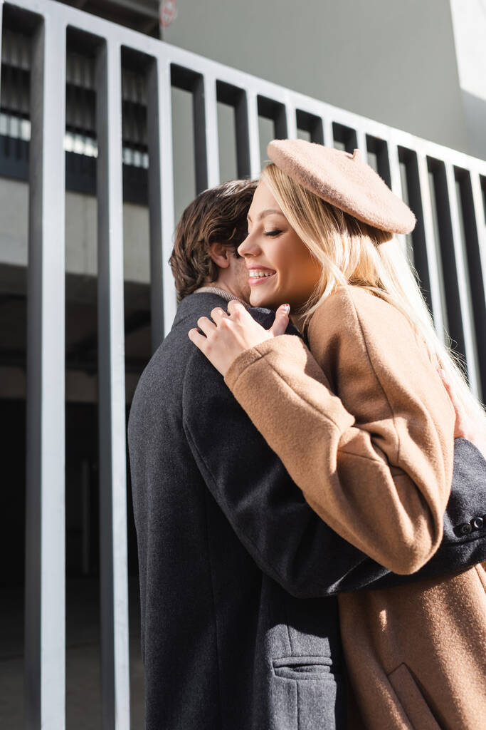 mulher alegre na boina abraçando com namorado perto de cerca na rua - Foto, Imagem