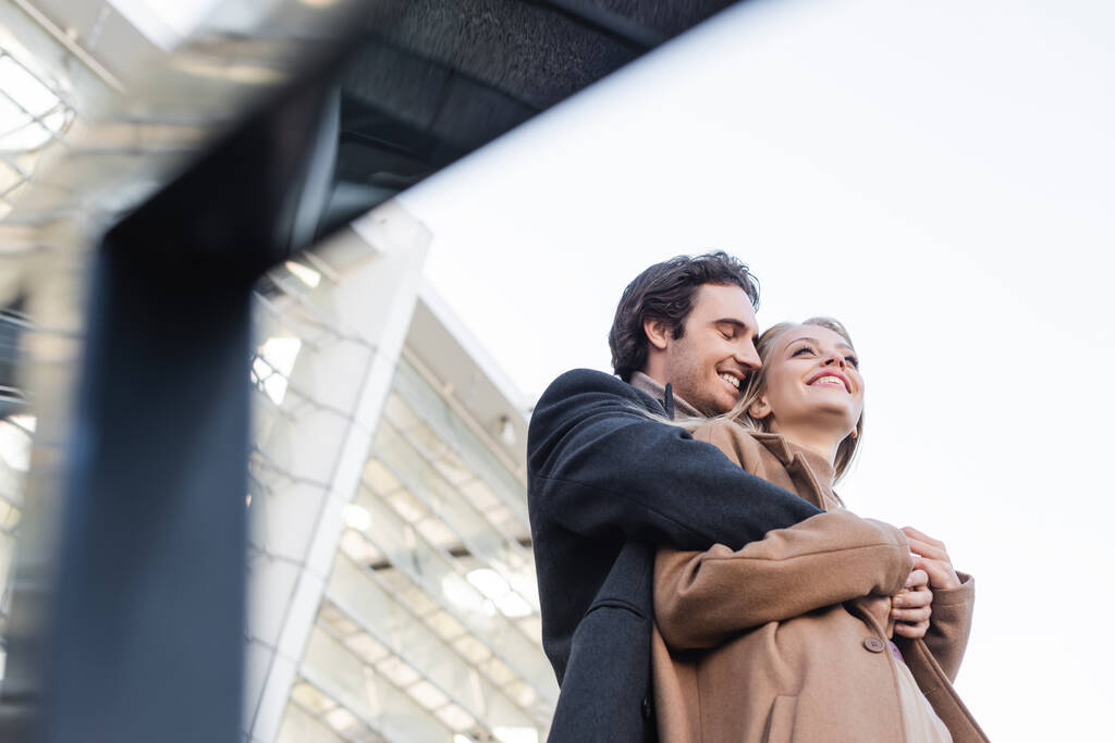 bajo ángulo vista de hombre en abrigo abrazando feliz novia en borrosa primer plano - Foto, imagen