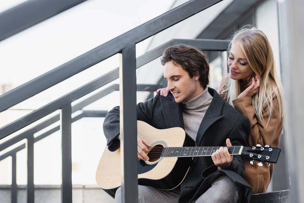 tevreden blonde vrouw aanraken schouder van vriendje spelen akoestische gitaar  - Foto, afbeelding