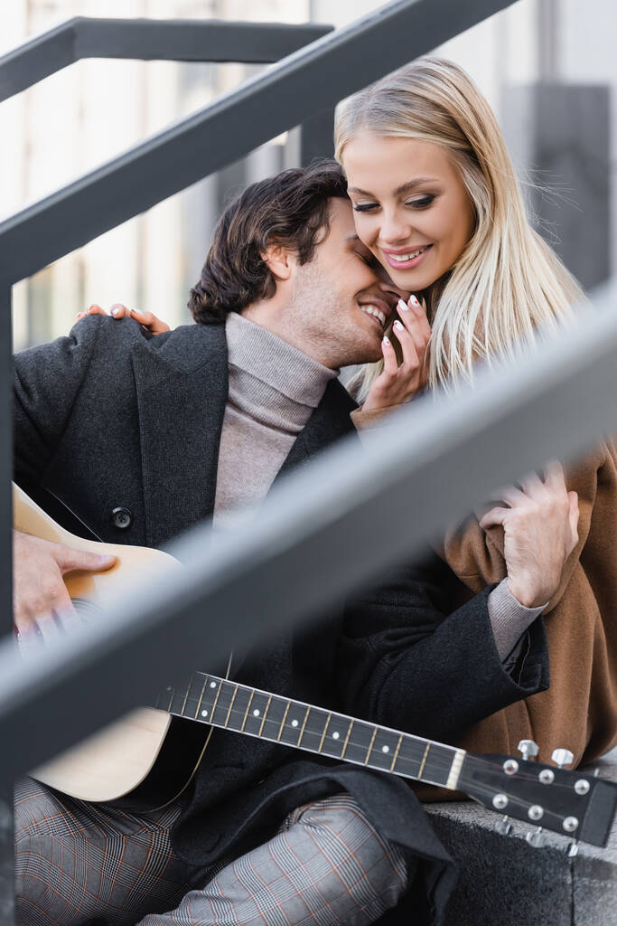 joven mujer rubia sonriendo cerca de hombre con guitarra acústica en primer plano borrosa - Foto, Imagen
