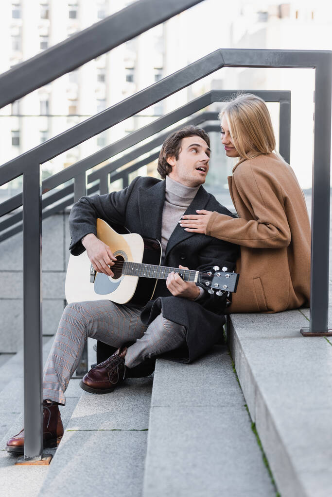 heureuse blonde femme en automne vêtements touchant homme jouant guitare acoustique sur les escaliers - Photo, image