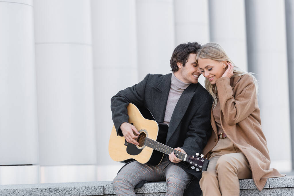 mężczyzna w jesiennym płaszczu gra na gitarze akustycznej w pobliżu szczęśliwej kobiety siedząc na parapecie - Zdjęcie, obraz