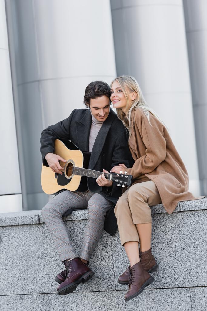 joyful woman in trendy autumn clothes sitting on parapet near man playing acoustic guitar - Photo, Image