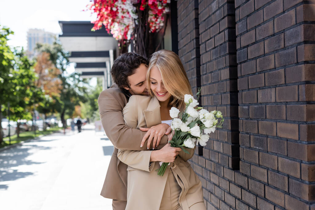 Neşeli bir kadın elinde bir buket Eustoma çiçeği tutarken erkek arkadaşı onu sokakta kucaklıyor. - Fotoğraf, Görsel