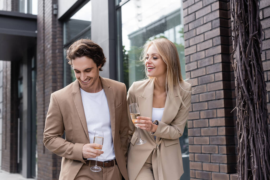 radostný mladý pár v béžových oblecích chůze s sklenicemi šampaňského na ulici - Fotografie, Obrázek