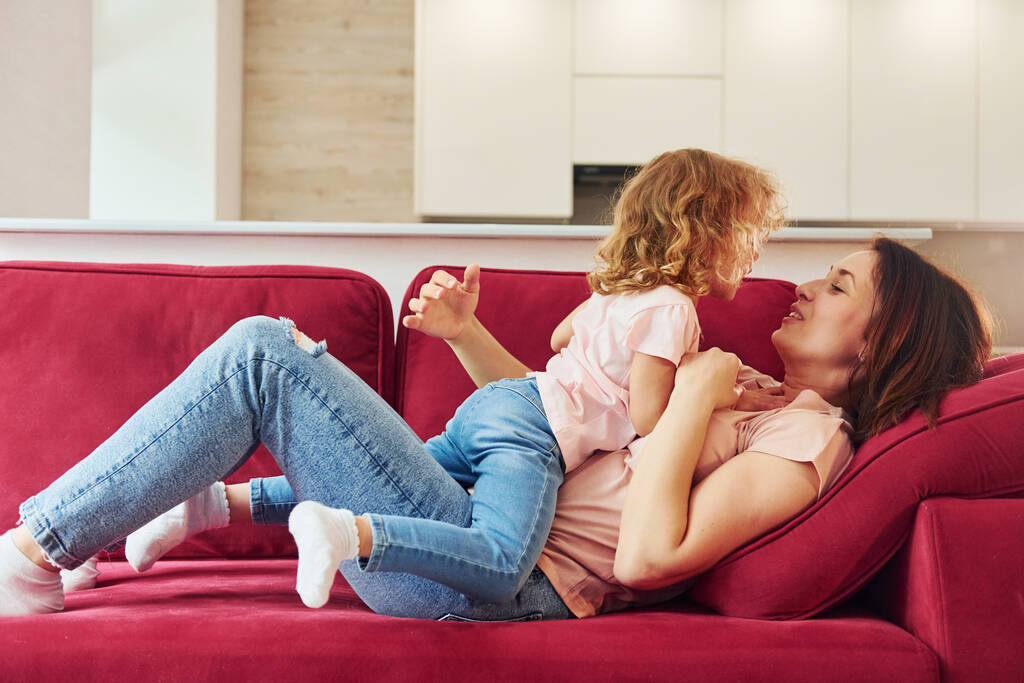Lying down on red sofa. Young mother with her little daughter in casual clothes together indoors at home. - Photo, Image