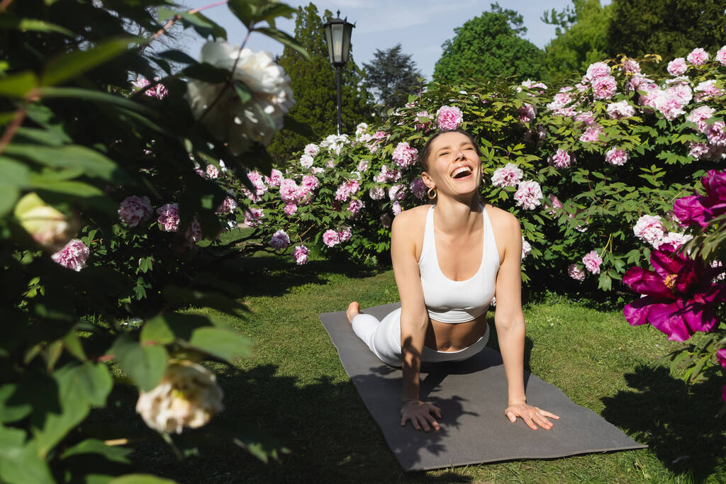smějící se žena se zavřenýma očima cvičící kobru pózující na podložce jógy poblíž kvetoucích rostlin - Fotografie, Obrázek