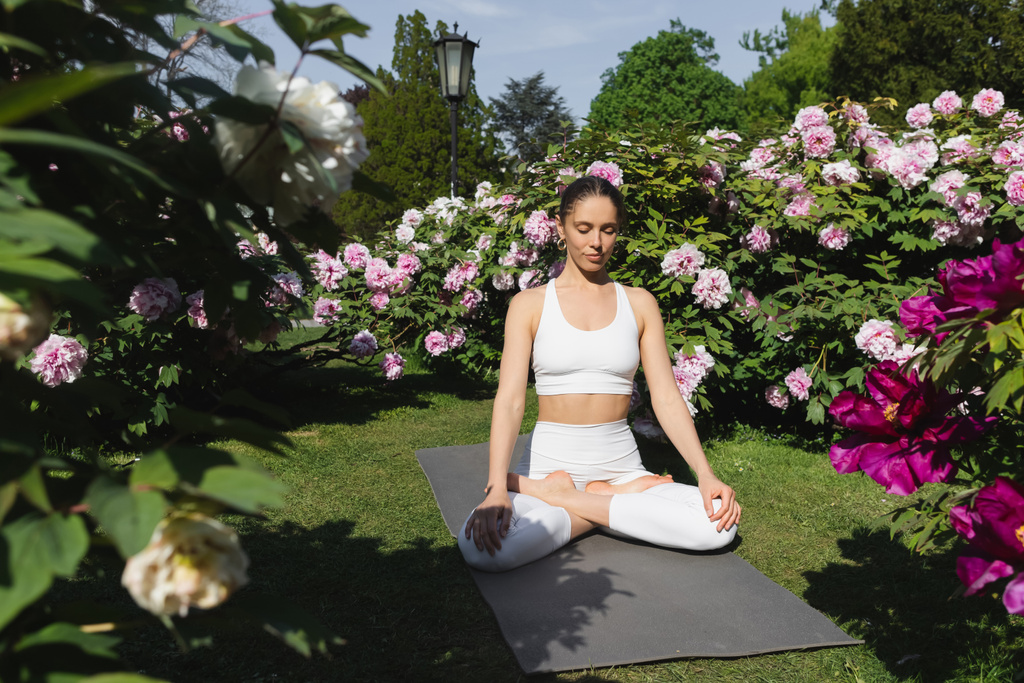 Frau mit geschlossenen Augen meditiert in Lotus-Pose neben blühenden Pflanzen im Park - Foto, Bild
