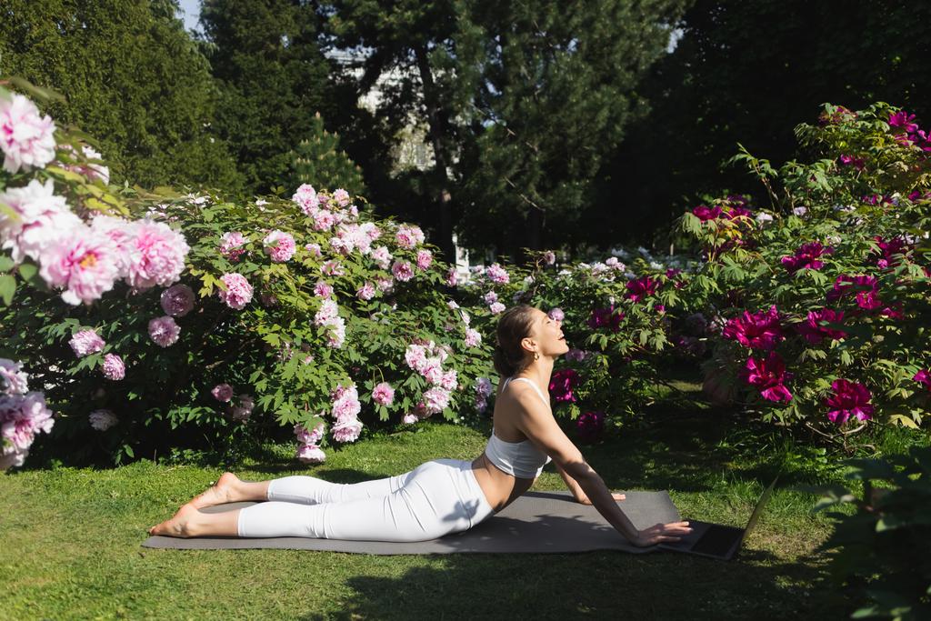side view of happy woman practicing yoga in cobra pose on lawn near laptop - Foto, imagen