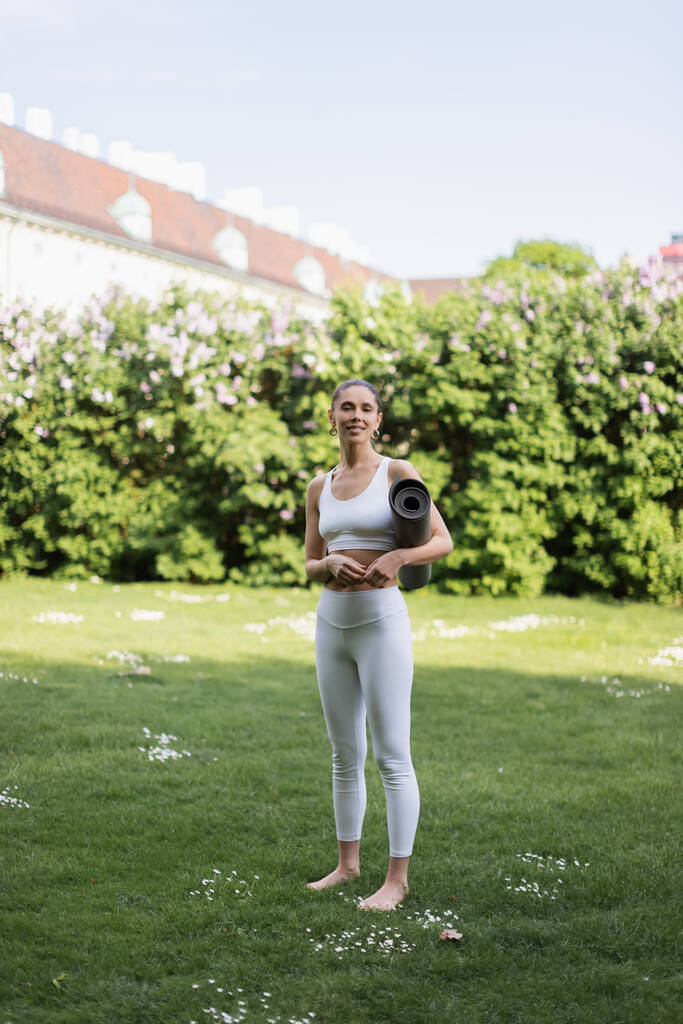 longitud completa de mujer descalza en ropa deportiva blanca de pie con estera de yoga en el césped verde - Foto, Imagen