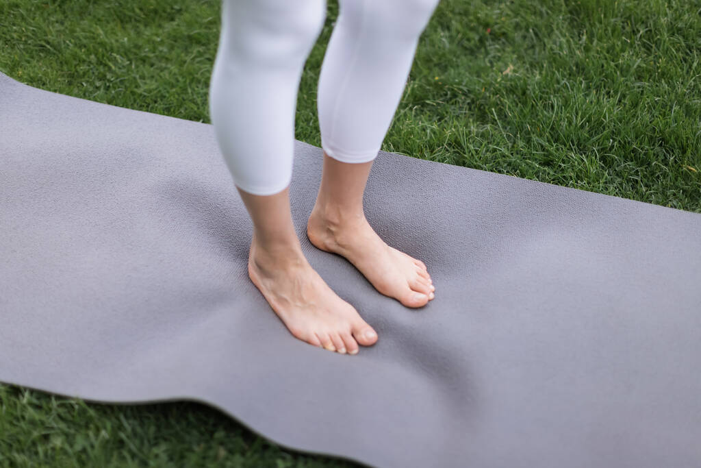 bijgesneden weergave van barefoot vrouw in wit leggings staande op yoga mat buiten - Foto, afbeelding