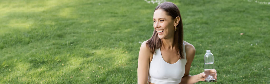 happy woman in white sports bra holding sports bottle and looking away outdoors, banner - Photo, image