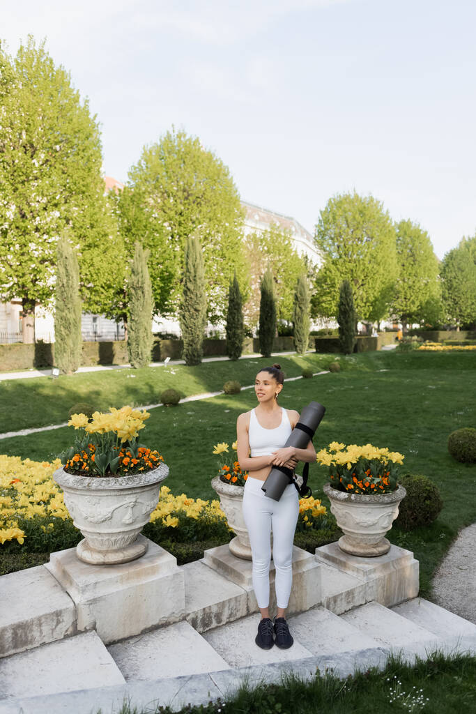 longueur totale de la femme en vêtements de sport blanc debout avec tapis de fitness près de vases de parc avec des fleurs - Photo, image