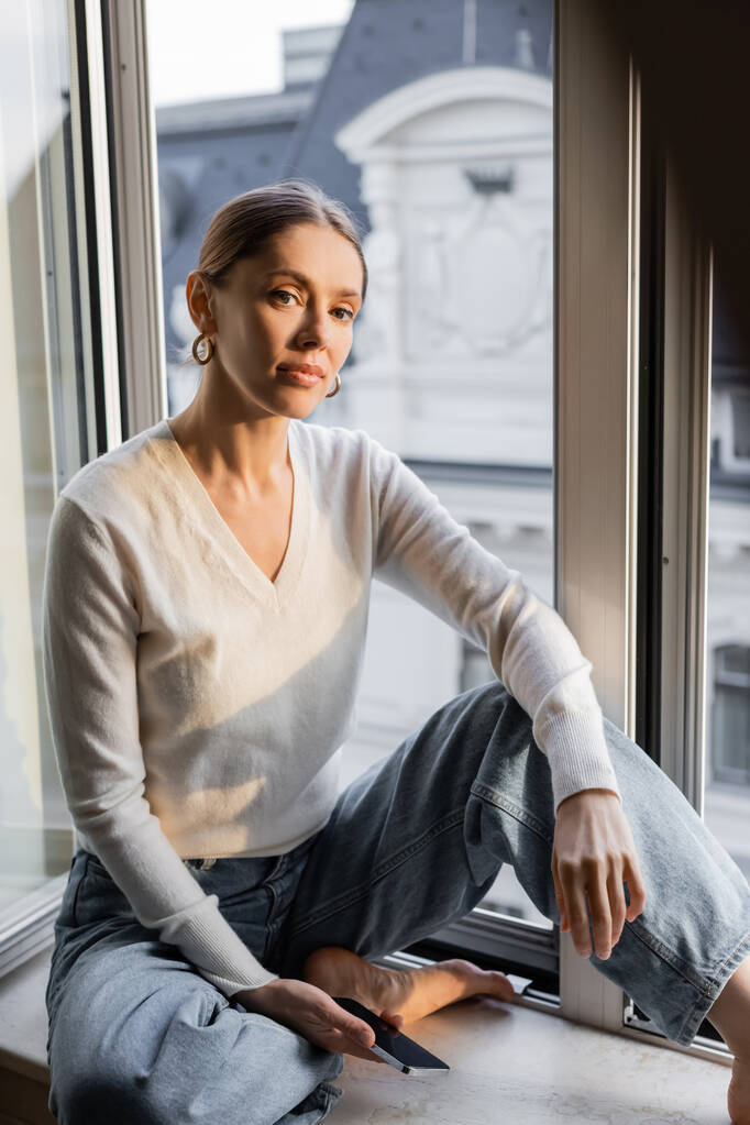 mezítlábas nő fehér pulcsiban ül az ablakpárkányon és a kamerába néz. - Fotó, kép