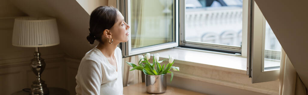 dreamy woman looking away through window near fresh tulips, banner - Foto, imagen
