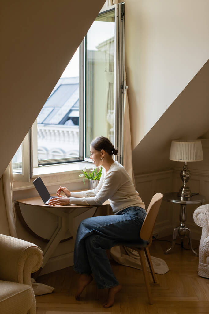 vue latérale de la femme tapant sur ordinateur portable dans une chambre mansardée confortable près de la fenêtre - Photo, image
