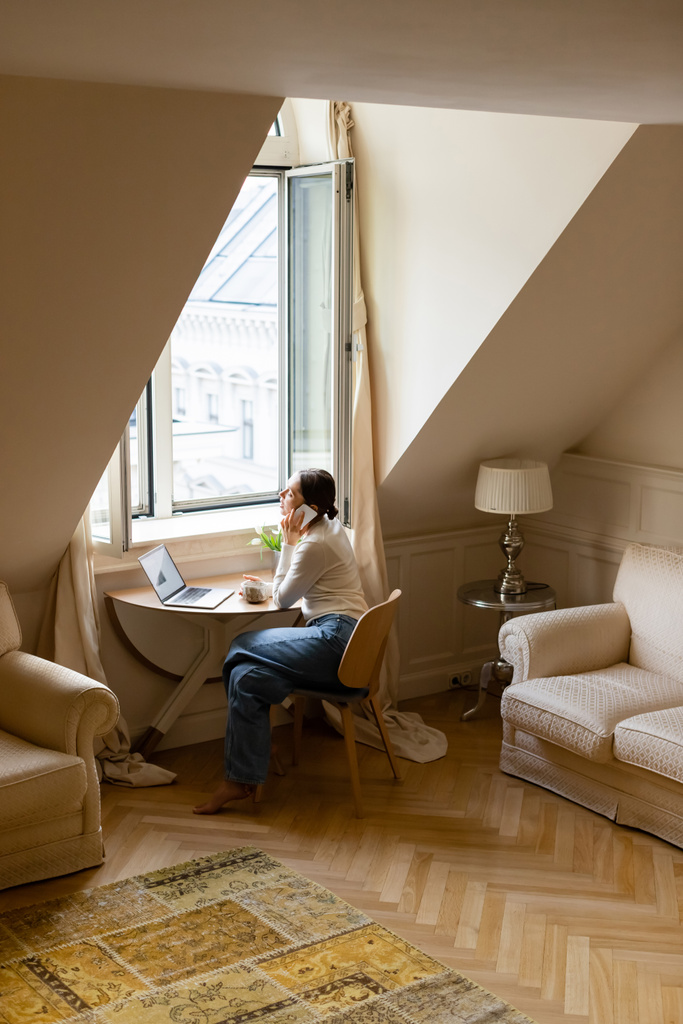 Босоногая женщина разговаривает по мобильному телефону возле ноутбука и окна в уютной мансарде - Фото, изображение