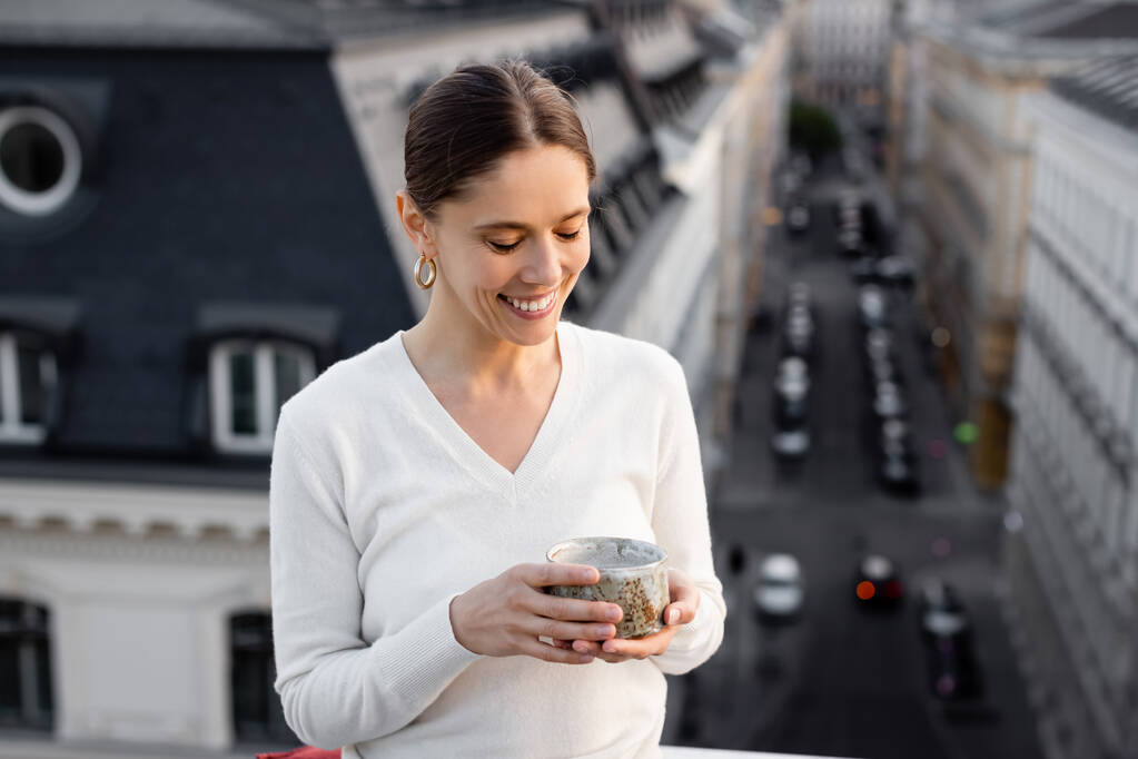 mujer alegre en jersey blanco sosteniendo taza de arcilla con té al aire libre - Foto, imagen