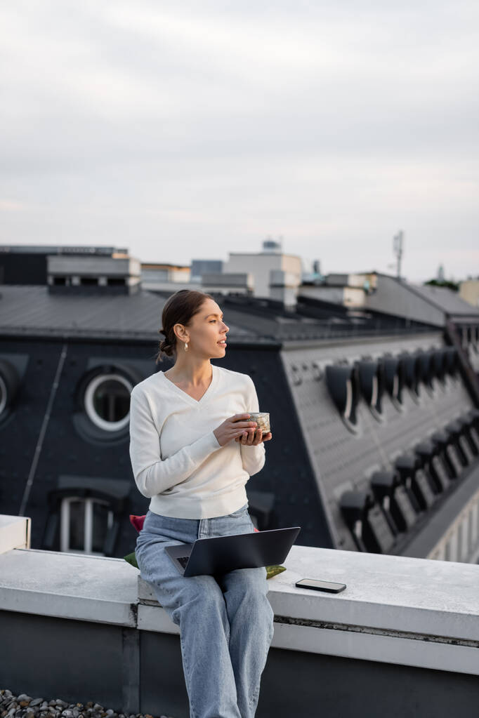 Kil bardaklı kadın laptopun ve cep telefonunun yanındaki çatıda oturuyor. - Fotoğraf, Görsel