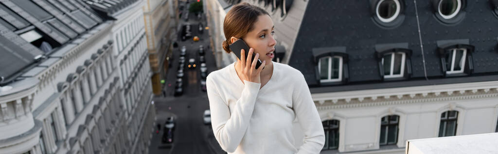 woman in white pullover talking on cellphone and looking away on rooftop, banner - Photo, Image