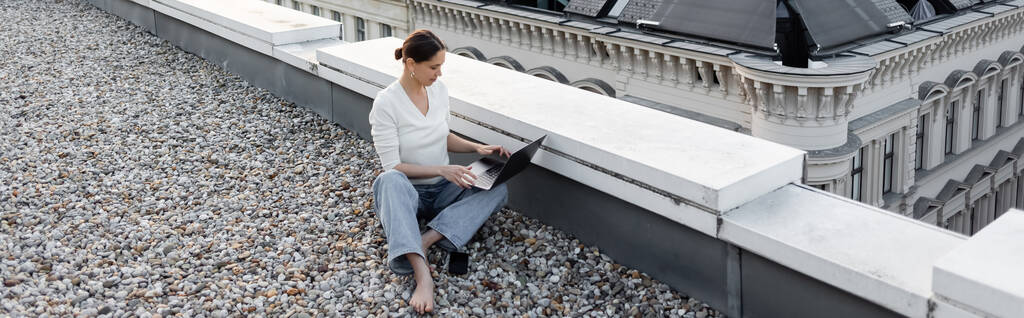 Hochwinkelaufnahme einer barfüßigen Frau, die am Laptop auf dem Dach arbeitet, Banner - Foto, Bild