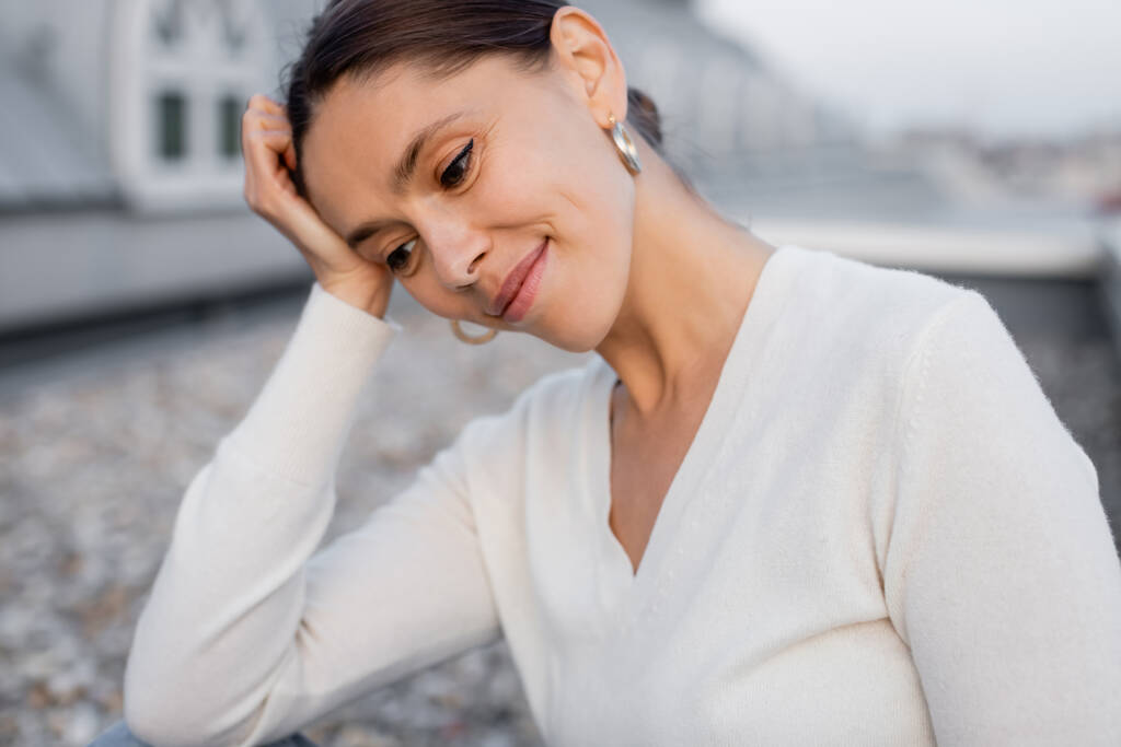 verträumte Frau im weißen Pullover, die die Hand vor dem Kopf hält und im Freien lächelt - Foto, Bild