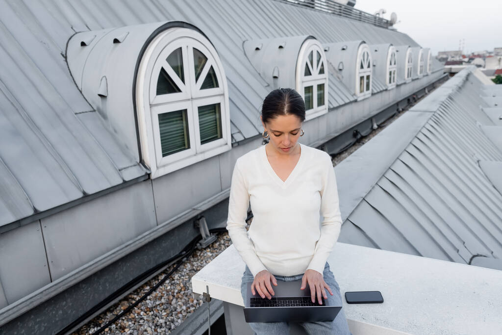 woman in white jumper typing on laptop near smartphone with blank screen on rooftop - Fotoğraf, Görsel