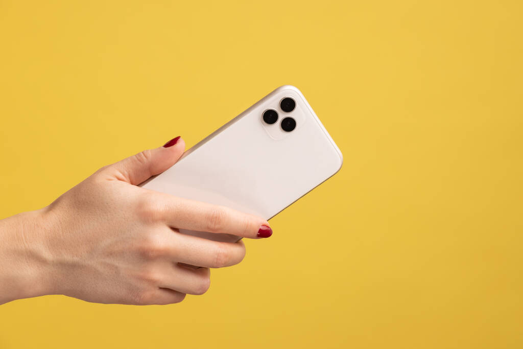 Retrato de perfil close-up da mão da mulher segurando o telefone celular, mostrando o lado de trás do telefone celular. Estúdio interior tiro isolado no fundo amarelo. - Foto, Imagem
