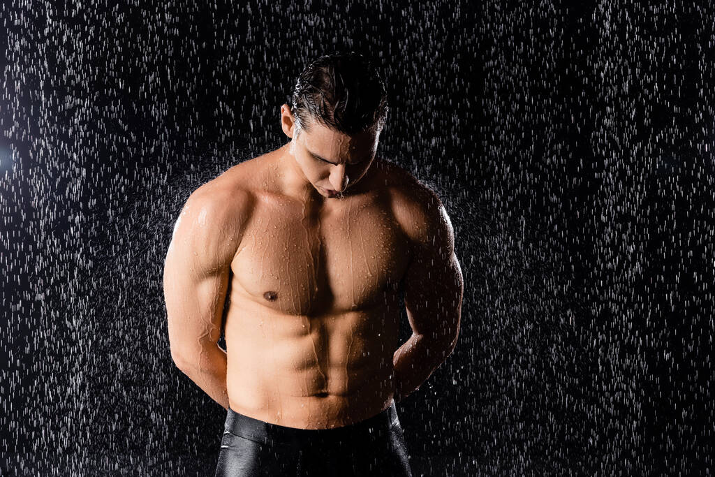 man met sterk lichaam staande onder regen met de handen achter de rug op zwarte achtergrond - Foto, afbeelding