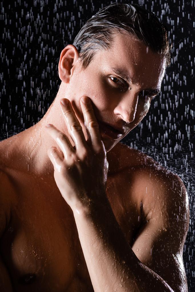 molhado homem tocando rosto e olhando para a câmera enquanto chuveiro no fundo preto - Foto, Imagem