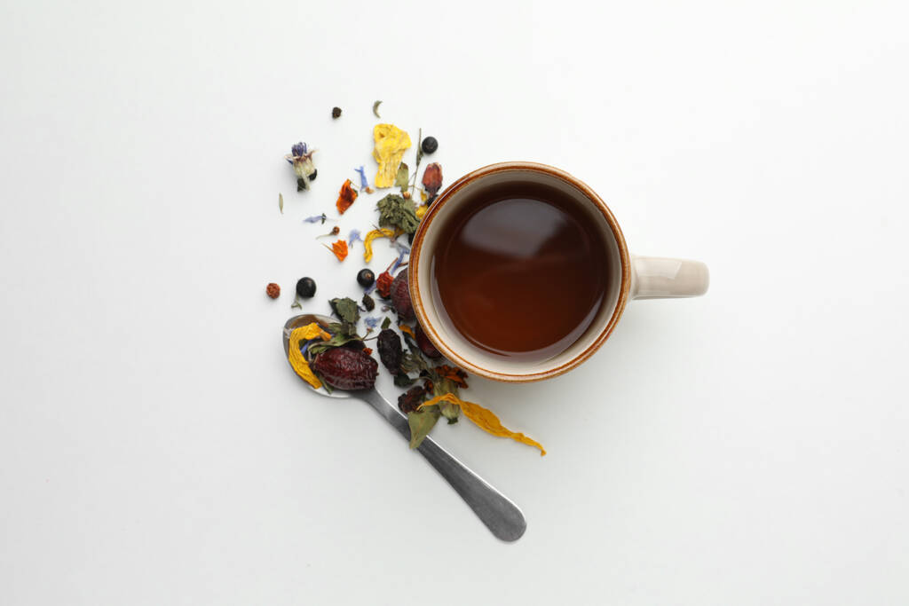 Cup of freshly brewed tea, dried herbs and spoon on white background, flat lay - Photo, Image