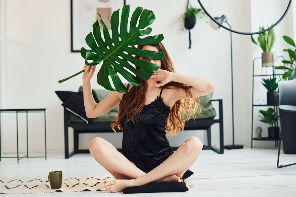 Young woman in pajamas sitting on the floor indoors at daytime and holding big green leaf. - Photo, Image