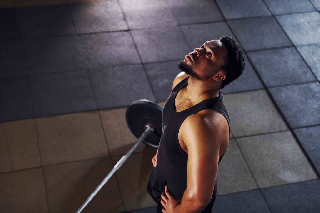 Vue de dessus. Barbell par terre. Homme afro-américain fort en vêtements de sport ont séance d'entraînement journée dans la salle de gym. - Photo, image
