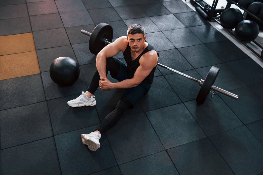 Starker Mann in sportlicher Kleidung sitzt mit Ausrüstung auf dem Boden im Fitnessstudio. - Foto, Bild
