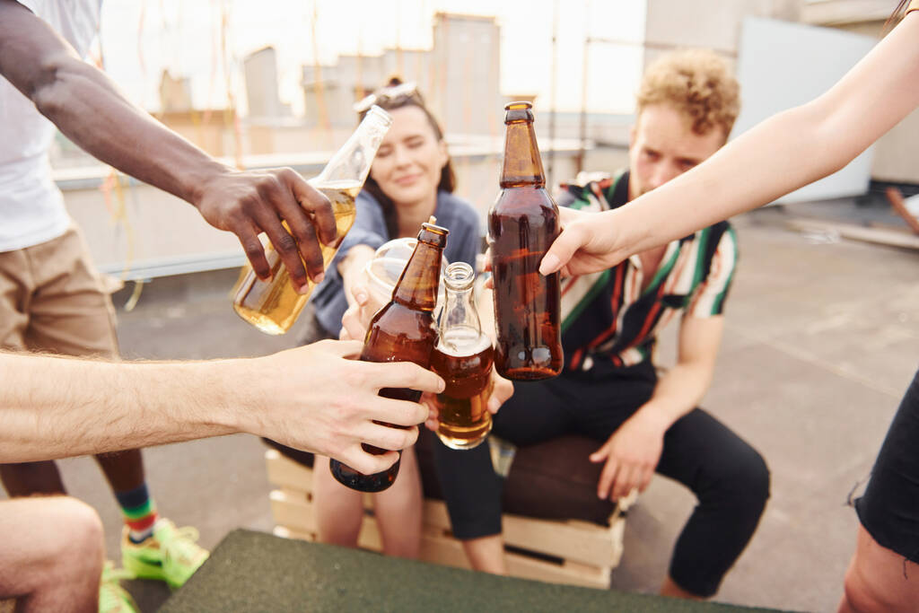Proost doen bij flessen met bier. Groep jongeren in casual kleding geven overdag samen een feestje op het dak. - Foto, afbeelding