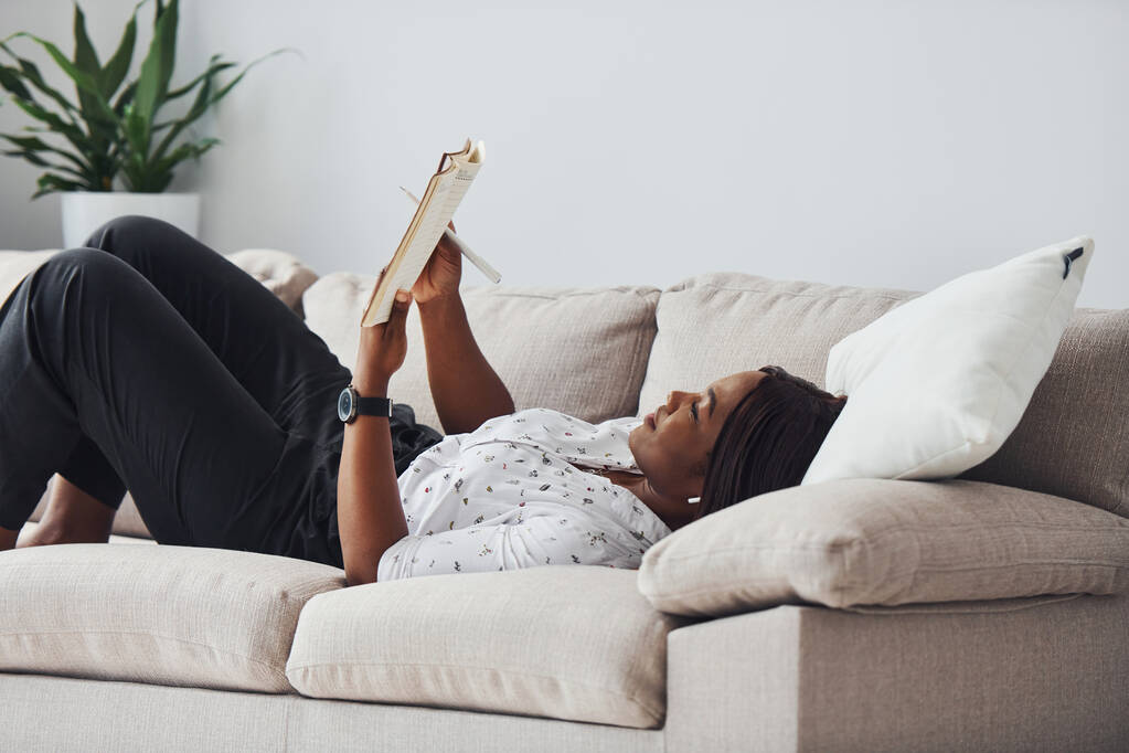 Mujer afroamericana positiva en camisa blanca acostada en sofá con bloc de notas en las manos. - Foto, imagen