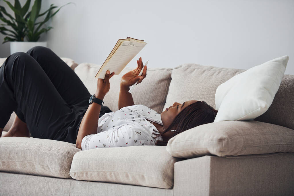 Femme afro-américaine positive en chemise blanche couchée sur le canapé avec un bloc-notes dans les mains. - Photo, image
