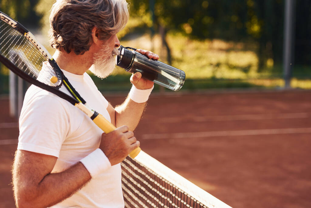 Pause machen und Wasser trinken. Stilvoller Senior in weißem Hemd und schwarzen Sporthose auf dem Tennisplatz. - Foto, Bild