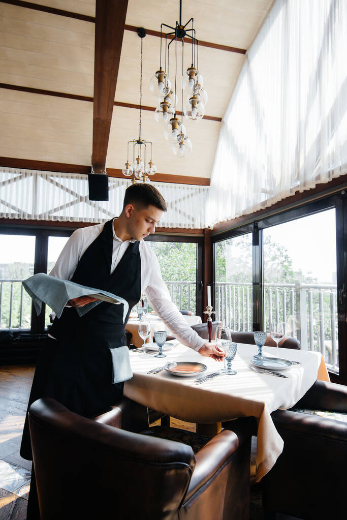 Un joven camarero con un elegante uniforme se dedica a servir la mesa en un hermoso restaurante gourmet. Actividad del restaurante, del más alto nivel. - Foto, imagen