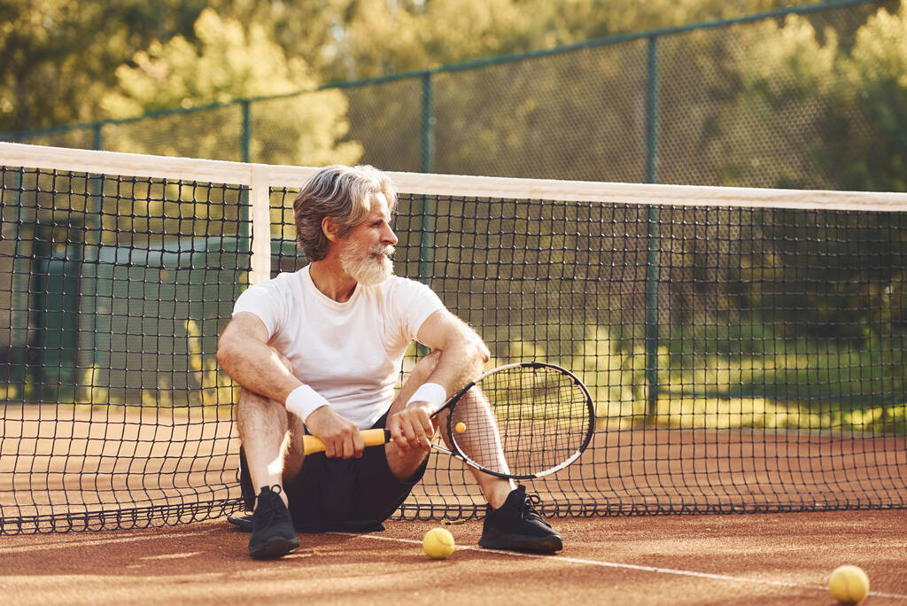 Сидіти і відпочивати. Старший стильний чоловік у білій сорочці та чорних спортивних шортах на тенісному корті
. - Фото, зображення