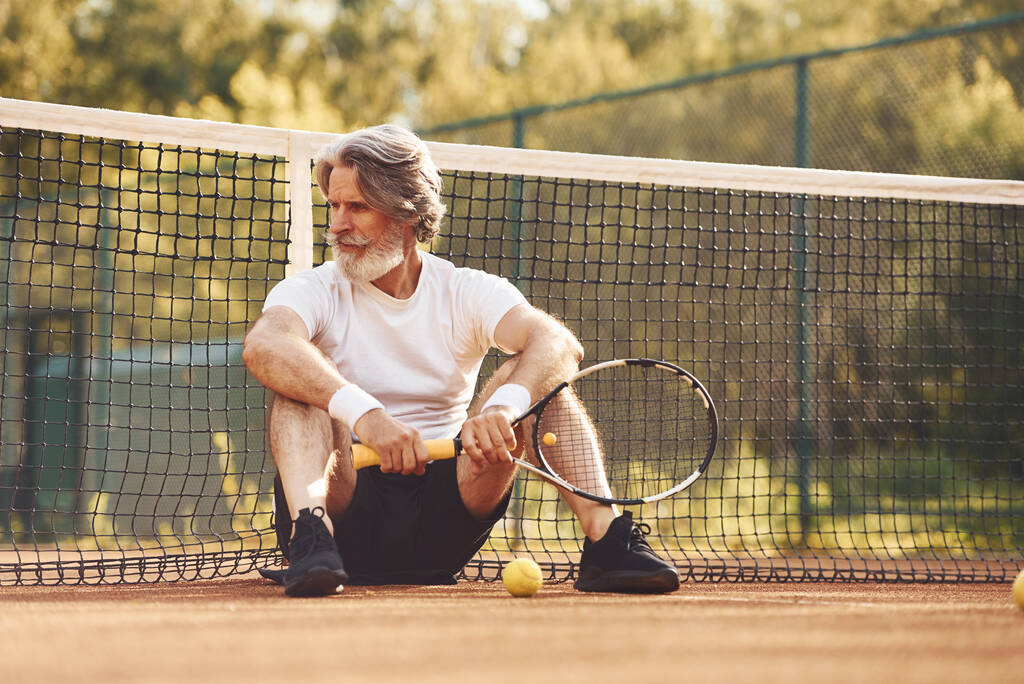 Ülök és pihenek. Senior stílusos férfi fehér ingben és fekete sportos rövidnadrágban a teniszpályán. - Fotó, kép