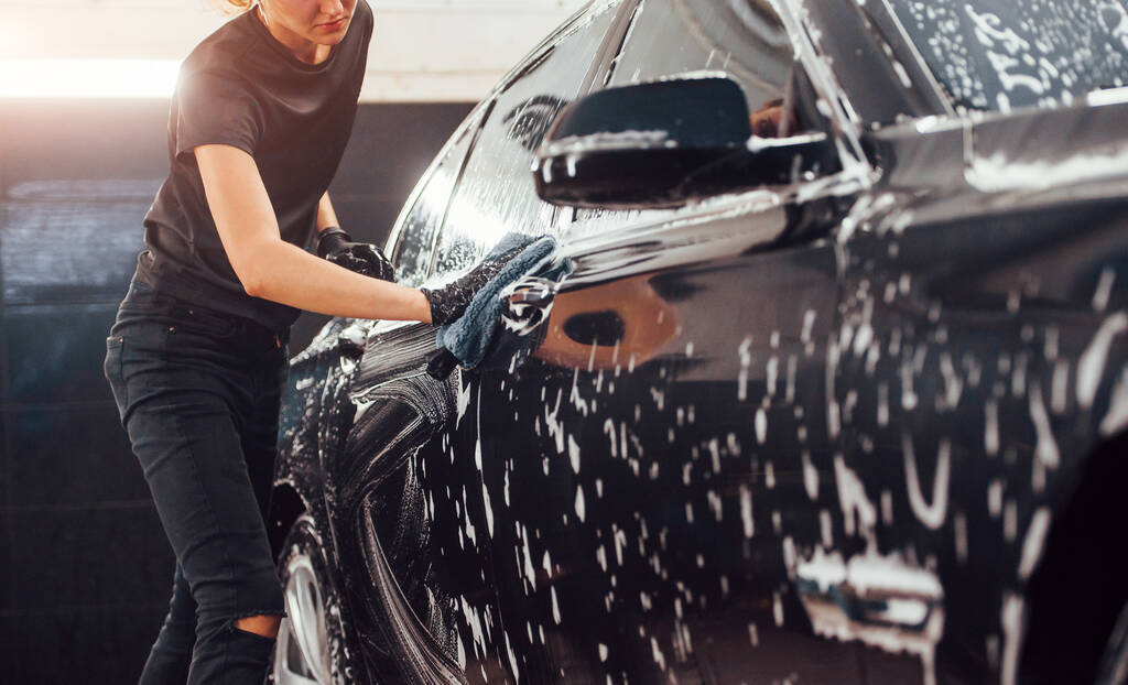 Wischt Fahrzeug, das in weißer Seife ist. Modernes schwarzes Auto wird von Frau in Waschanlage gereinigt. - Foto, Bild