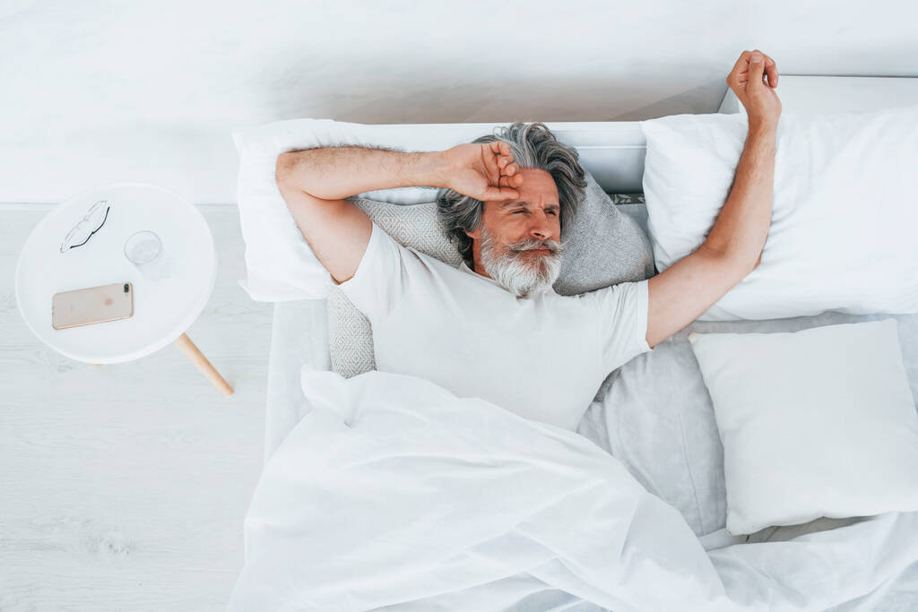 Homme moderne élégant senior avec cheveux gris et barbe à l'intérieur couché sur le lit et se réveiller. - Photo, image