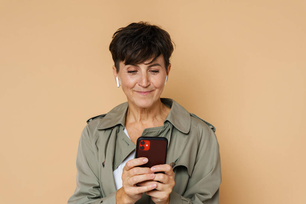 Mature european woman in earphones laughing and using cellphone isolated over beige background - Photo, Image