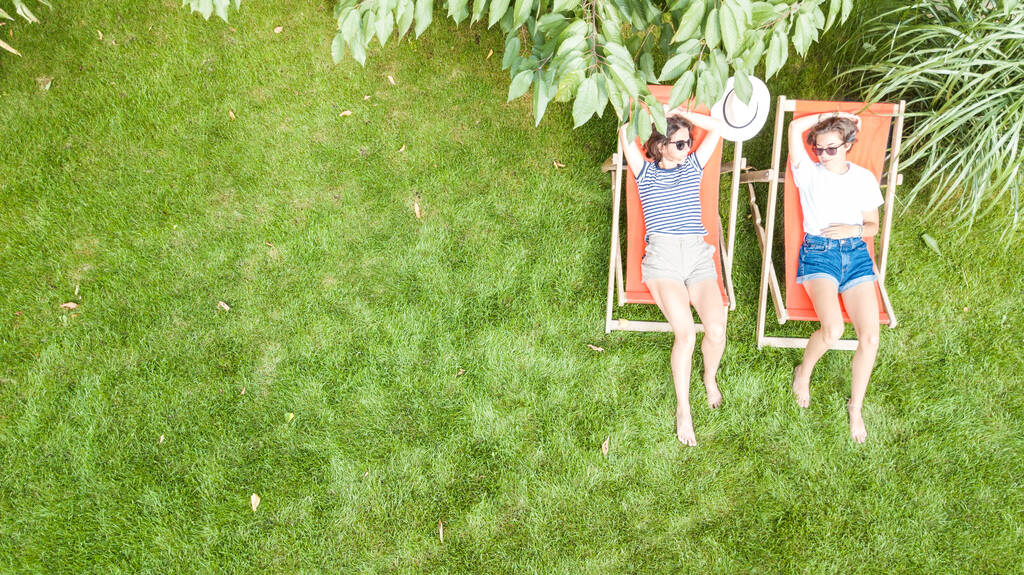 Молоді дівчата відпочивають в літньому саду на шезлонгах на траві, жінки друзі розважаються на відкритому повітрі в зеленому парку на вихідних, вид зверху на повітря
 - Фото, зображення