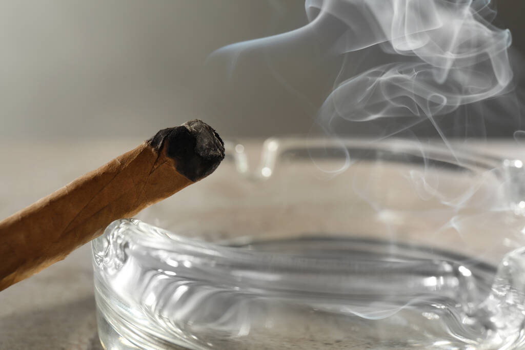 Smoldering sigaar in de buurt van glas asbak op lichtgrijze tafel, close-up - Foto, afbeelding